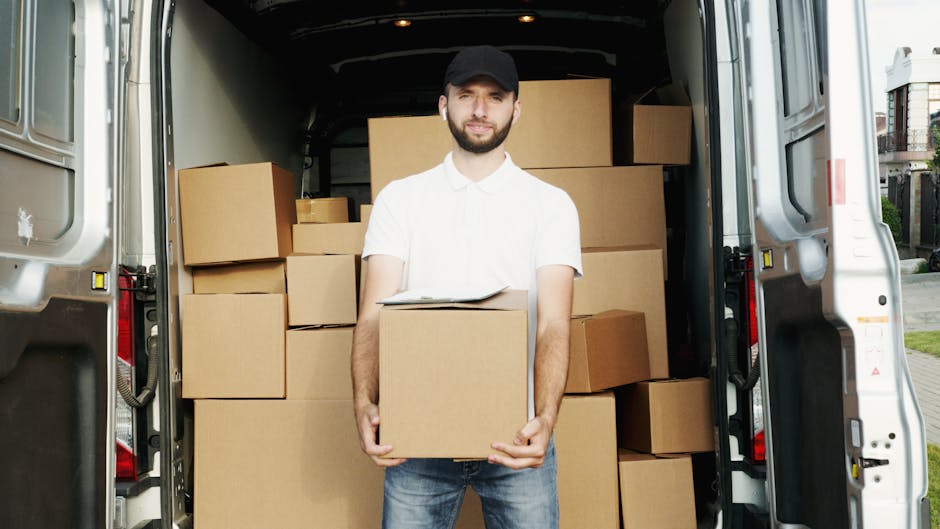 A man with a beard, wearing a white t-shirt and jeans, is standing on the pavement beside a light grey moving van with its side door open, engaged in loading or unloading cardboard boxes. The boxes va