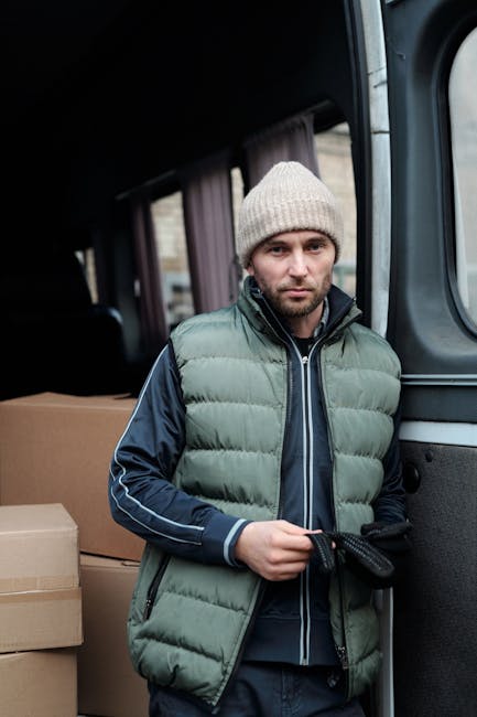 A young man with a beard and wearing a black cap and white polo shirt stands inside a moving van with the rear doors open during a home relocation. Behind him, the van is filled with stacked cardboard