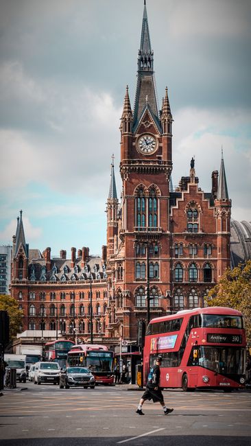 Photograph taken outdoors in an urban setting featuring a large, historic red brick building with ornate architectural details, towers, and a central clock. In the foreground, a busy street scene incl