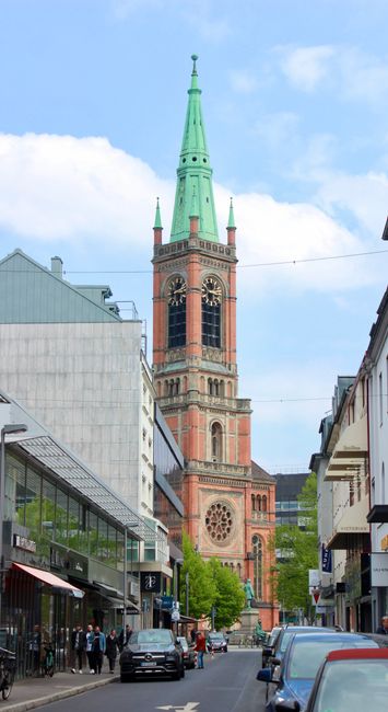 A street scene in St Johns Wood with a tall, historic church tower featuring a green spire and brick detailing visible against a partly cloudy sky. Modern retail shops with glass storefronts and awnings line both sides of the street, with parked cars along the pavement. Several cardboard boxes, wrapped furniture, and packing materials are situated on the sidewalk near a van, indicating ongoing home relocation or furniture transport. The loading process appears to involve individuals moving household items, including large pieces of furniture covered with protective blankets and strapped for secure handling. A trolley or dolly is also visible nearby, assisting with the transportation of items from the property to the vehicle. The scene captures elements of packing, loading, and street-level logistics typical of professional removals, supported by [COMPANY_NAME], in the context of house removals and relocation services on NW8, with a focus on access tips and logistical planning for high street moves.
