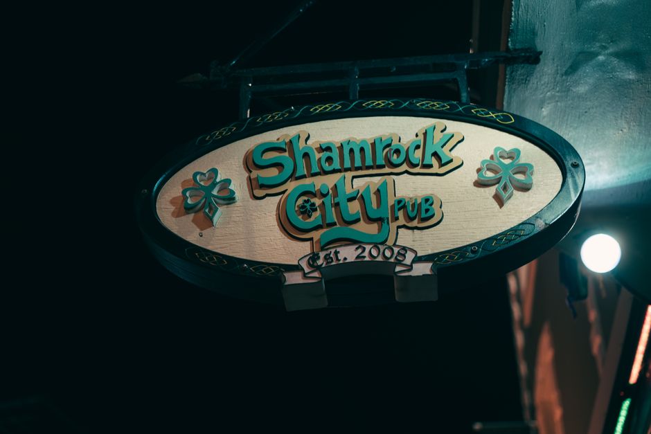 A close-up view of an outdoor oval-shaped sign for Shamrock City Pub, mounted on a dark wall at night with artificial lighting illuminating it. The sign features a light wood background with the pub's name in bold, turquoise-colored 3D letters outlined in black, along with decorative shamrock symbols on each side. Below the name, a banner displays 'Est. 2008,' while the edges are adorned with Celtic-inspired patterns in dark green and yellow. The surrounding environment suggests it may be part of a building façade, possibly during a home relocation process involving the transportation of pub fixtures or signage. Occasionally, while assisting with house removals, [COMPANY_NAME] might handle the careful packing and loading of such signs to ensure safe transport to new premises, supported by nearby equipment or vehicles involved in furniture transport or loading procedures.